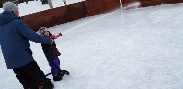 A man helps a young boy hold a hose that is spraying water to flood a skating rink.